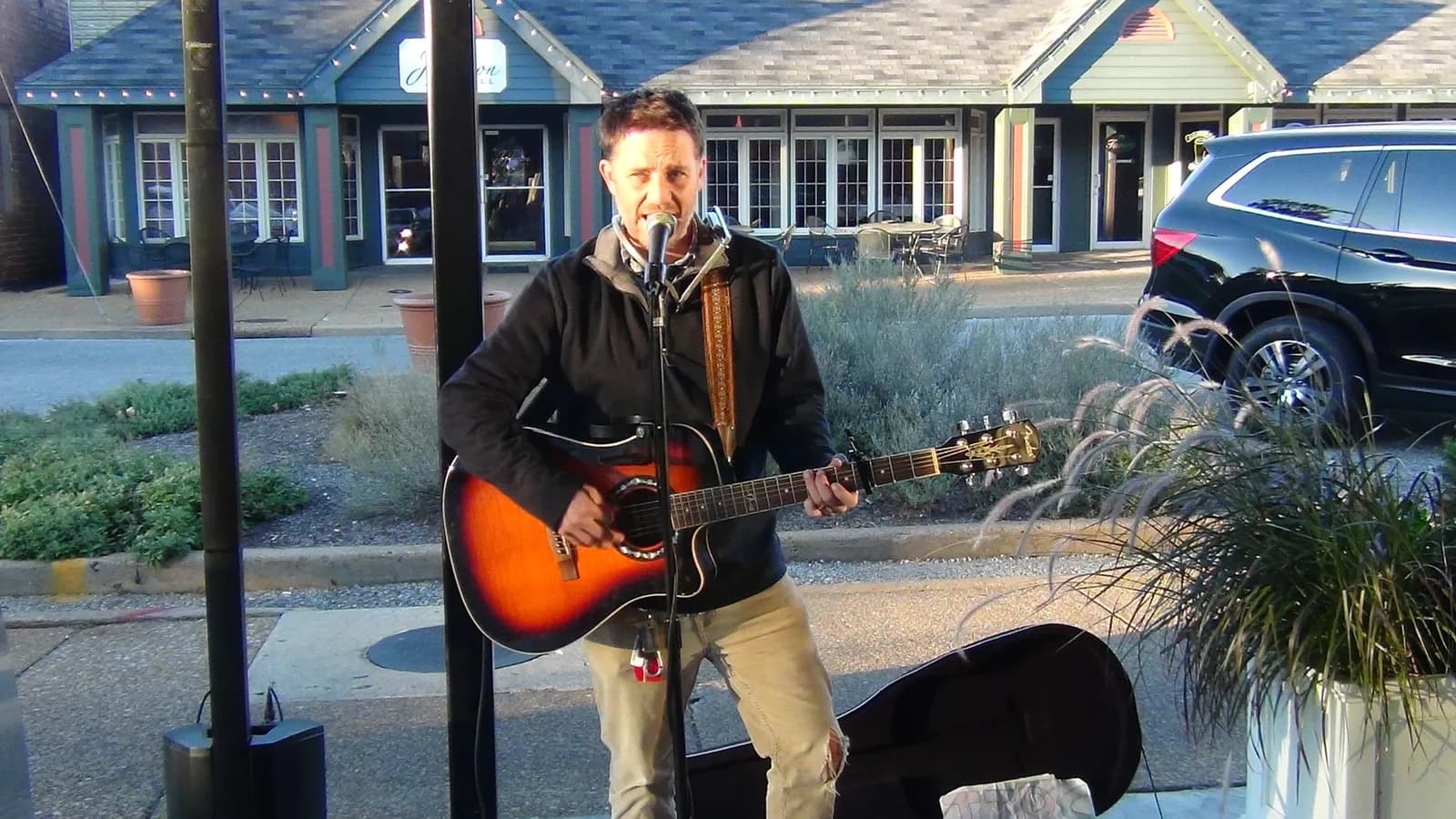 A guitarist singing into a microphone on a patio outside a row of storefronts, an open guitar case at his feet.