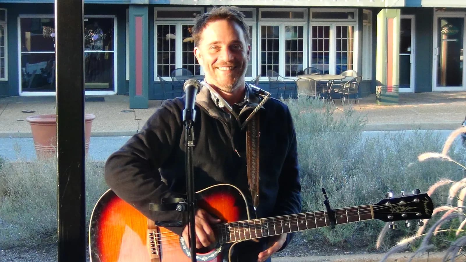 A guitarist standing at a microphone outside a row of storefronts, smiling toward the camera.