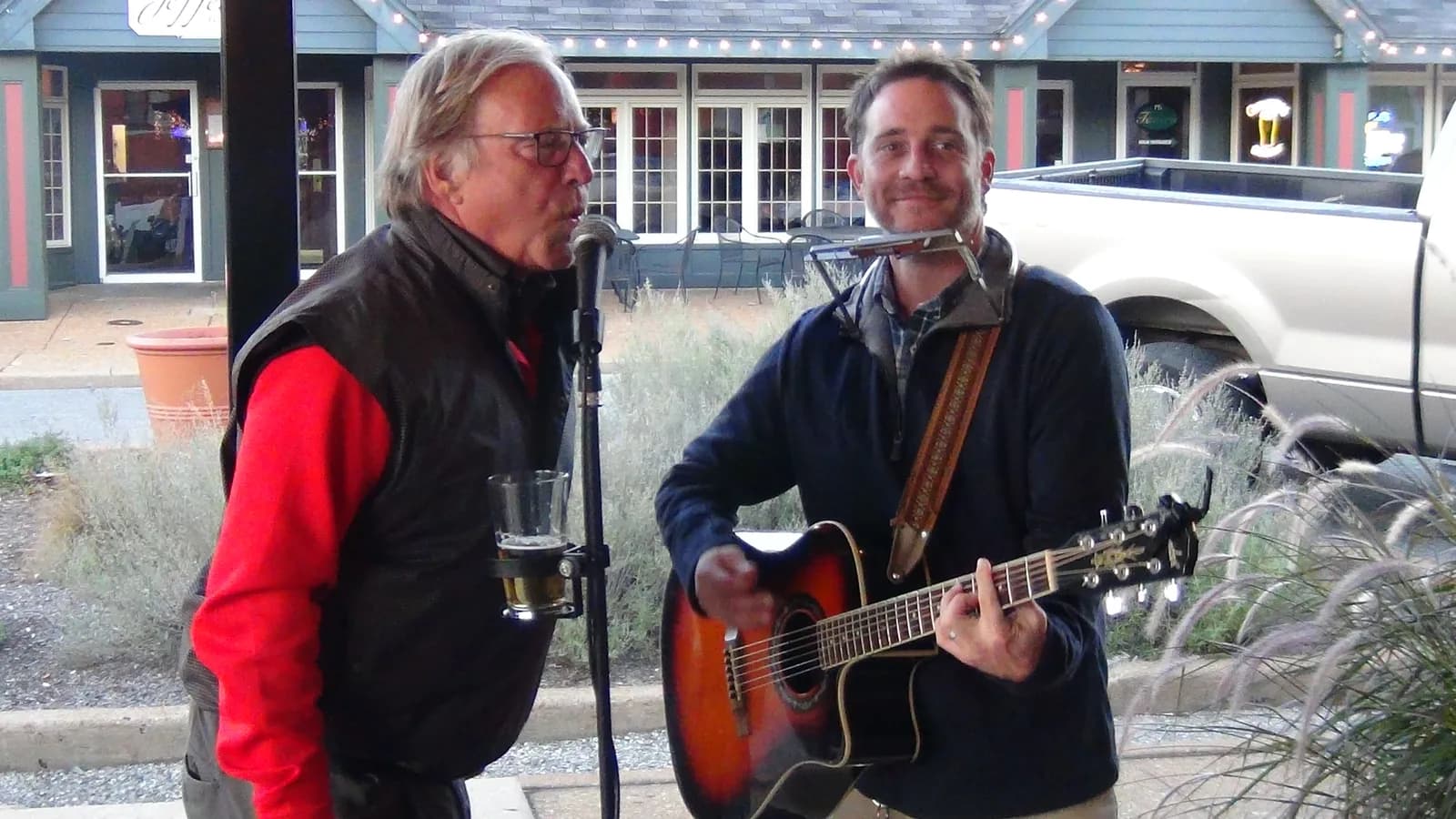 Two musicians sharing a microphone outside a row of storefronts, one playing guitar with a harmonica holder around his neck.
