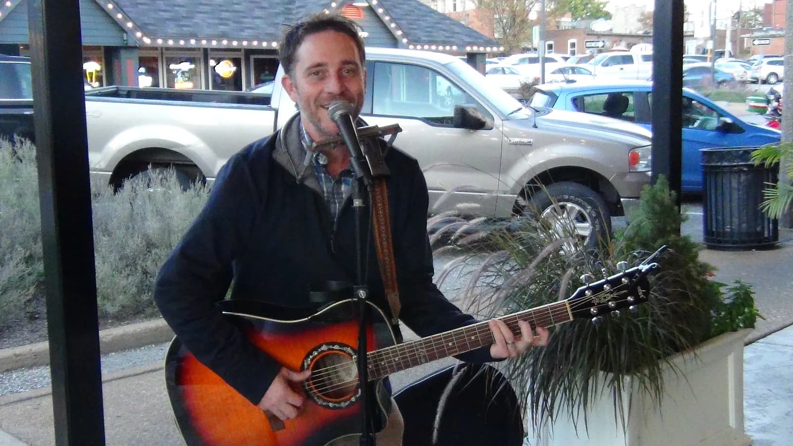 A guitarist singing into a microphone on a patio, with a parking lot behind him.