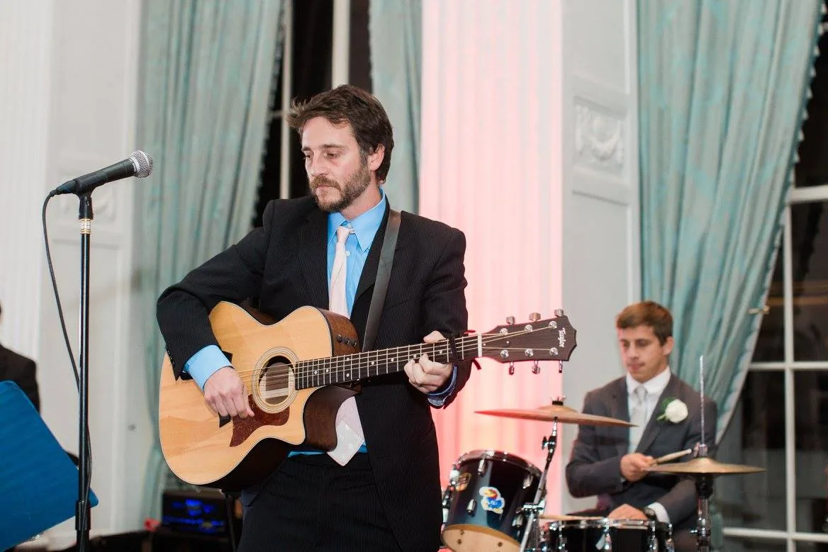 Chris Cahill playing guitar in a suit, with the band’s drummer at the kit behind him at an indoor event.
